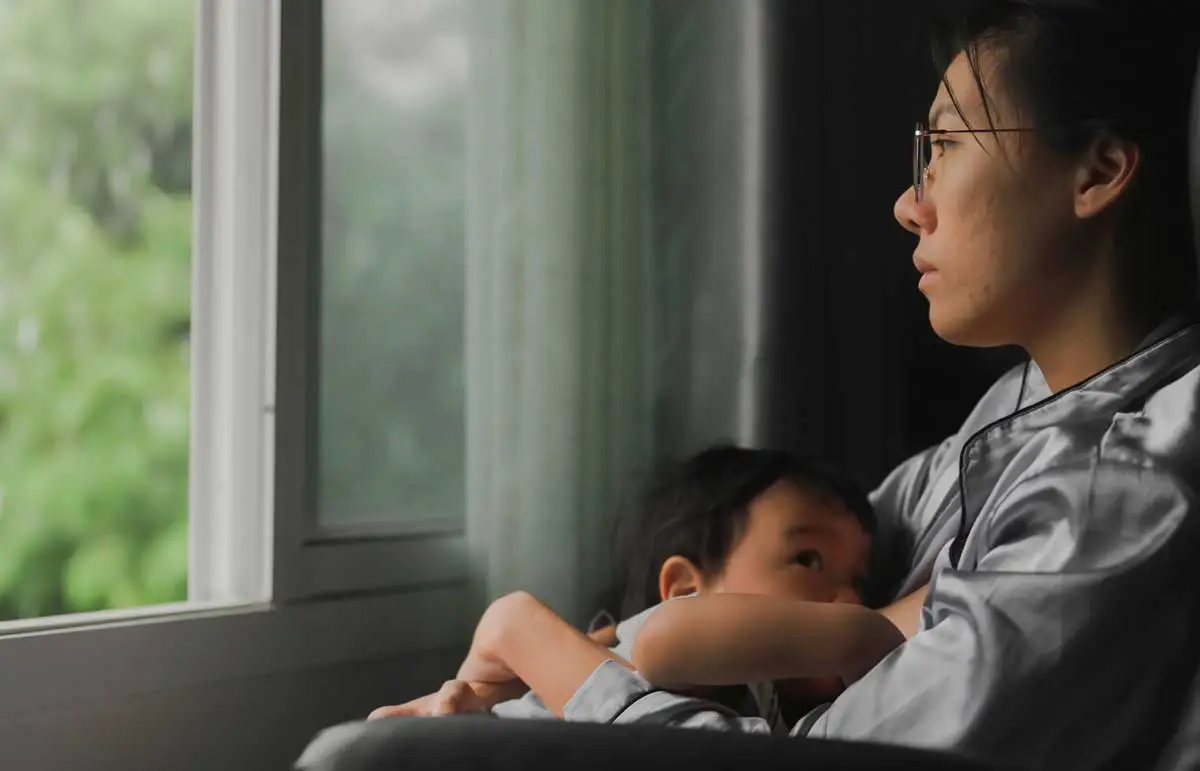 Tired mother breastfeeding her baby while looking out of a grey window.